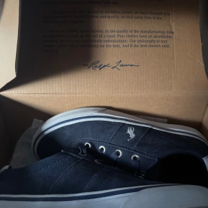 Mörkblå sneakers från Ralph Lauren - Snygga mörkblå sneakers från Ralph Lauren med vit logga broderad på sidan. Skorna har ovandel i mocka, vita detaljer vid hälen och vit sula med blå rand. Klassisk snörning och metallöljetter ger en stilren look. Perfekta för dig som gillar en clean och sportig stil.