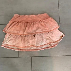 Rosa volangkjol från Superdry - Supersöt kort volangkjol från Superdry i ljusrosa bomull. Kjolen har två lager volanger och resår i midjan för en bekväm passform. Broderad Superdry-logga i guld på nederkanten