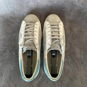 Philippe Model sneakers i mocka - Snygga sneakers från Philippe Model i ljusgrå mocka och vitt skinn med blågrön detalj på hälen. Klassisk låg modell med snörning och platt sula. Perfekta för dig som gillar stilrena och trendiga skor med en twist.