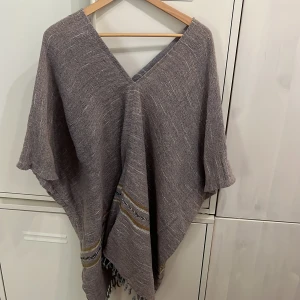 Grå stickad poncho - Poncho i grå stickat bomull och linne material med v-ringad hals och lösa, vida ärmar. Nedtill finns dekorativa ränder i gult, svart och vitt samt fransar längs kanten. 