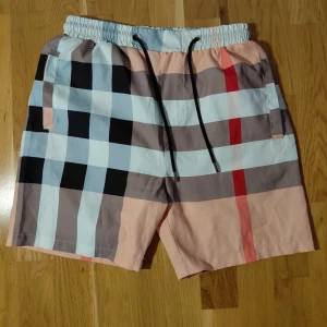 Bad byxor Burberry shorts  - Badshorts från Burberry med klassiskt rutigt mönster i beige, svart, vit, röd. Modellen har elastisk midja med svart snörning och sidofickor. Använd men har inga eventuella skador eller fläckar. Storlek S