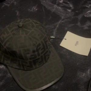 Svart keps med Fendi-logga - Snygg svart keps från Fendi med klassiskt FF-monogram över hela kepsen. Kepsen har böjd skärm och justerbar rem baktill. Tillverkad i bomull och har en exklusiv känsla med tydlig Fendi-tag på insidan. Perfekt för dig som vill sticka ut med en lyxig accessoar.