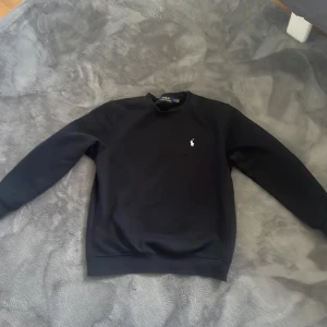 Svart tröja från Polo Ralph Lauren - Klassisk svart tröja från Polo Ralph Lauren med rund hals och diskret broderad logga på bröstet. Tröjan har lång ärm och är tillverkad i mjukt material som passar perfekt till en stilren look.