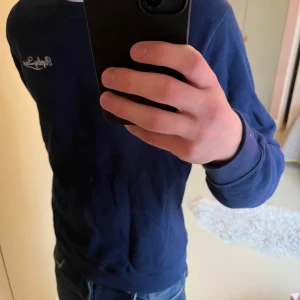 Marinblå sweatshirt från Replay - Säljer en väldigt snygg marinblå sweatshirt från Replay med broderad vit Replay League-text på bröstet. Tröjan har rund hals och är gjord i mjuk bomull, perfekt för kyligare dagar. Enkel design som funkar till jeans.