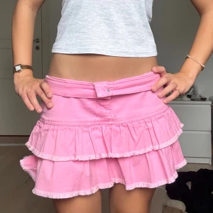 Rosa volangkjol från Tommy Hilfiger - Supersöt rosa kortkjol från Tommy Hilfiger med två lager volanger och råa kanter. Kjolen har bälteshällor, knapp och dragkedja framtill samt en metallogo med T och stjärnor bak. Perfekt för dig som vill sticka ut med en färgglad och lekfull stil.