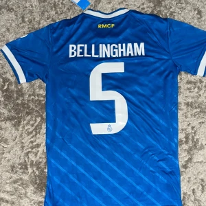 Real Madrid Bellingham tröja blå Adidas M - Snygg blå Real Madrid fotbollströja från Adidas med Bellingham och nummer 5 på ryggen. Tröjan har vita detaljer, klubbmärke, sponsortryck och klassiska Adidas-ränder på axlarna. Tillverkad i lätt och ventilerande material, perfekt för match eller träning.
