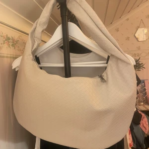 Beige axelväska med flätad look - Stor beige axelväska med snygg flätad struktur. Väskan har dragkedja upptill och ett rymligt innerfack med extra ficka. Perfekt för dig som vill ha plats för allt du behöver under dagen och gillar stilren design. Väskan är helt oanvänd. 