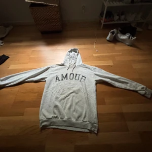 Grå hoodie med tryck 'AMOUR' - Grå hoodie med svart text 'AMOUR' på bröstet och en stjärna tryckt på ryggen. Tröjan har huva med snörning och långa ärmar. Perfekt för chill dagar och enkel att matcha med jeans eller joggers.