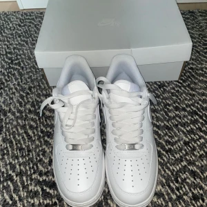 Nike Air Force 1 helvita sneakers - Klassiska Nike Air Force 1 sneakers i helvitt skinn med perforerad tå, rund tå och platt sula. Skorna har snörning och diskret Nike-logga på sidan. Perfekta för dig som vill ha en clean och tidlös look. Levereras med originalkartong.