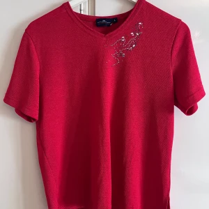 Röd topp med strass från Navigazione - Röd topp från Navigazione med v-ringning och korta ärmar. Toppen har dekorativa strasstenar och små silvriga detaljer vid axeln som ger en snygg touch. Materialet är mjukt och lättstickat, perfekt för dig som gillar färg och lite extra bling.