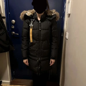 Svart lång dunjacka Parajumpers XS - Säljer en svart lång dunjacka från Parajumpers med päls på huvan och ikonisk gul rem vid axeln. Jackan har dragkedja framtill, flera fickor och patch på ärmen. Perfekt för kalla vinterdagar och riktigt snygg passform. använt den endast under en vinter. Den har inte kemtvättas, så priset kan diskuteras vid snabb affär. Jackan är i bra skick och är i storlek XXS.