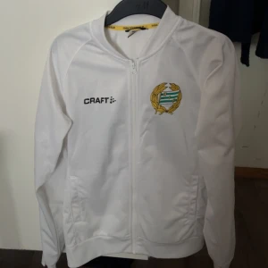 Vit Hammarby Craft ziptröja S - Vit ziptröja från Craft med Hammarby IF:s emblem på bröstet och stort grönt 'BAJEN' tryckt på ryggen. Tröjan har hel dragkedja, ståkrage och två fickor framtill. Perfekt för dig som vill visa ditt stöd för Bajen.