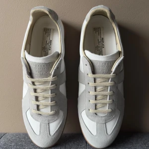 Maison Margiela sneakers vit/grå - Maison Margiela sneakers i vitt och ljusgrått med ovandel i skinn och mocka, klassisk gummisula och snörning. Snygg retrostil med rund tå och diskreta detaljer. Perfekta för dig som gillar minimalistisk och stilren design.                      Lägg prisförslag om du har ett pris i tanke