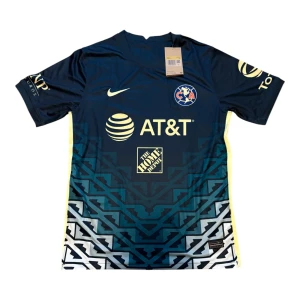 Club América matchtröja Nike blå - Snygg Club América fotbollströja från Nike i mörkblått med gula detaljer och grafiskt mönster nertill. Tröjan har klubbmärke på bröstet, flera sponsorer och är tillverkad i lätt, ventilerande polyester. Perfekt för dig som vill sticka ut på planen eller läktaren.