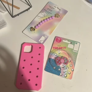 Rosa mobilskal med LED Pop-Deco och handrem - Snyggt rosa mobilskal med hålmönster, komplett med LED Pop-Deco (rosettformad dekoration) och lila pärlhandrem med gulddetaljer. Perfekt för att ge din mobil en personlig och lekfull stil. Alla delar är nya och oanvända i förpackning.iphone13