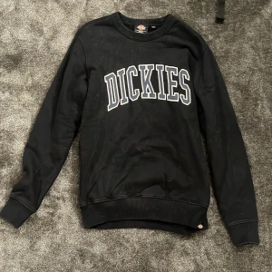 Svart sweatshirt från Dickies - Svart sweatshirt från Dickies i storlek small med stort vitt och grått DICKIES-tryck på bröstet. Tröjan har rund hals, ribbade muddar och är långärmad. Perfekt för en chill och avslappnad stil. Materialet är mjukt och känns skönt mot huden.