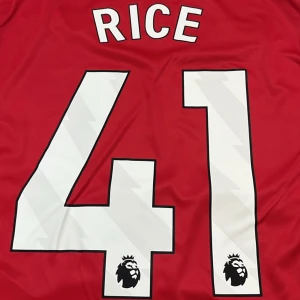 Arsenal fotbollströja Rice 41 Adidas - Snygg Arsenal fotbollströja från Adidas med Rice och nummer 41 på ryggen och en till med Gyökeres . Framsidan har Emirates Fly Better-tryck och klubbens klassiska kanonlogga. Tröjan är vit framtill med subtilt mönster och röda detaljer, ryggen är röd med stora vita siffror. Tillverkad i lätt och ventilerande material.