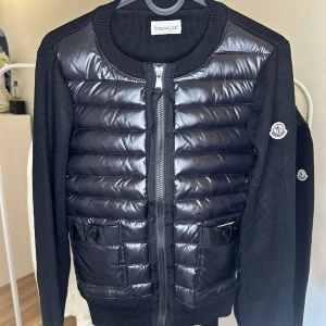 Moncler Cardigan - Svart pufferjacka från Moncler med glansig, quiltad front och stickade ärmar. Jackan har rund hals, dragkedja och två fickor med knappstängning. Moncler-logga på ärmen ger extra stilpoäng. Perfekt för dig som vill ha en snygg och modern look.