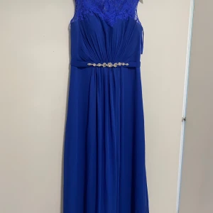 Blå långklänning med spets och strass - Elegant blå långklänning med spets upptill och ärmlös design. Klänningen har vackra draperingar framtill och ett bälte med strassdetaljer i midjan som ger extra lyxig känsla. Perfekt för dig som vill sticka ut med en klassisk och stilren look. Det finns dessutom en body och en sjal till. 