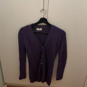 Lila kofta från Dagmar med knappar - Snygg lila kofta från Dagmar med smal passform och v-ringning. Koftan har långa ärmar och stängs med tre knappar framtill. Materialet är stickat och har en diskret struktur. Perfekt för lager-på-lager och att styla med jeans eller kjol.