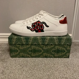 Gucci vita sneakers med orm och ränder - Gucci sneakers i vitt skinn med klassiska gröna och röda ränder på sidorna. Ena sidan har en broderad röd och svart orm. Baktill är ena skon rödmetallic och den andra svartmetallic. Platt sula och snörning framtill. Riktigt snygga och unika detaljer.