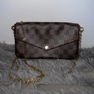 Louis Vuitton Pochette Félicie väska - Snygg Louis Vuitton Felicie axelväska i klassiskt brunt Damier Ebene-mönster med guldfärgad kedja och kuvertformad front. Insidan är fodrad i rött tyg och har en praktisk innerficka. Perfekt för dig som vill ha en ikonisk och trendig designerbag. Köptes för 8500 kr som visas på kvittot, idag värd 14 400 kr. Knappen är utbytt och ny! Väskan är knappt använd! Orginalförpackning och kvitto finns kvar. 