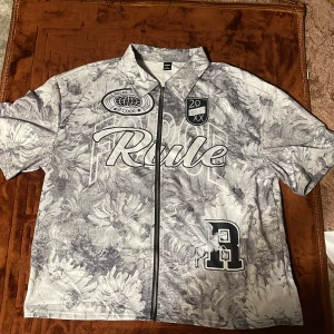 Blommig zip-tshirt från MANFINITY - Snygg t-shirt med blommigt gråvitt mönster från MANFINITY. Stora tryck med texten 'Rule', emblem och Pegasus-tryck framtill. Dragkedja hela vägen, klassisk krage och korta ärmar. Perfekt för dig som vill sticka ut med streetstyle-vibe.