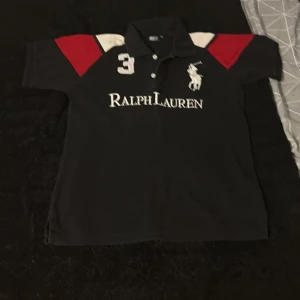 Svart Polo Ralph Lauren t-shirt M - Svart t-shirt från Polo Ralph Lauren med vita och röda detaljer på axlarna. Stor logga och text framtill, samt nummer 3 broderat. Klassisk pikékrage och korta ärmar. Tillverkad i bomull för en skön och sportig känsla.