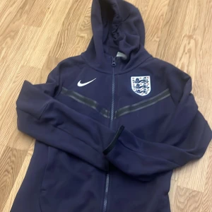 Mörkblå England hoodie från Nike - Snygg mörkblå hoodie från Nike med England-emblem på bröstet och vit swoosh. Tröjan har huva, hel dragkedja och långa ärmar. Perfekt för dig som gillar fotboll och vill visa support för England. Materialet känns mjukt och sportigt.
