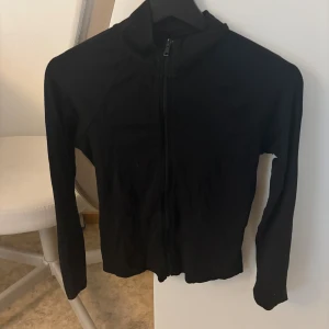 Svart ziptröja från Gina Tricot - Svart långärmad ziptröja från Gina Tricot i storlek M. Tröjan har hel dragkedja framtill och en enkel, stilren design med hög krage. Perfekt att slänga på sig över ett linne eller t-shirt. Materialet är mjukt och stretchigt för en bekväm passform. Liten i storleken!