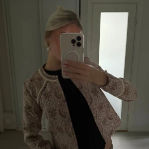 Paisleymönstrad kavaj från Boohoo - Paisleymönstrad kavaj i beige och brunt från Boohoo Boutique. Kort, rak modell med rundad halsringning och dekorativa kantband. Perfekt för dig som vill sticka ut med en unik och trendig jacka.