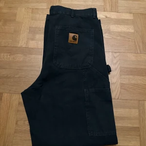 Svarta Carhartt cargopants loose fit - Svarta cargopants från Carhartt i loose original fit. Byxorna har flera stora fickor, hammarögla på sidan och klassisk Carhartt-logga på bakfickan. Tillverkade i 100% bomull för en slitstark känsla. Perfekta för dig som gillar streetwear och funktionella detaljer.