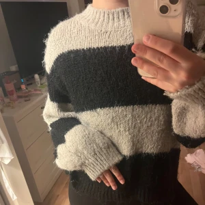 Randig stickad tröja i svart och grått - Mysig stickad tröja med breda svarta och ljusgrå ränder. Tröjan har en rund halsringning, ribbade muddar och lång ärm. Perfekt för dig som gillar en chill och avslappnad stil.