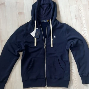 Marinblå zip up från Polo Ralph Lauren - Snygg marinblå hoodie från Polo Ralph Lauren med vit broderad logga på bröstet. Tröjan har dragkedja, huva med vita snören och två stora fickor framtill. Insidan är mjuk och fleecefodrad för extra komfort. Perfekt för en chill och stilren look. Storlek L men tycker att den sitter som M
