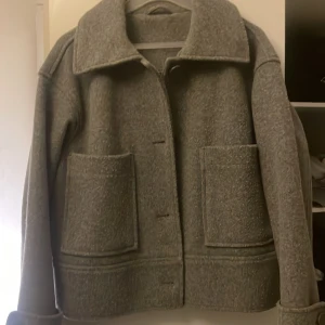 Grå ulljacka med stora fickor - Säljer en grå jacka i ull med stora framfickor och klassisk krage. Jackan har knäppning med stora knappar 