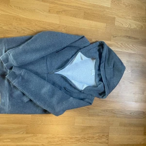 Grå hoodie med dragkedja från Mango - Grå hoodie från Mango i storlek M. Tröjan har huva med snörning, dragkedja framtill och två fickor. Tillverkad i mjuk bomullsblandning, perfekt för kyliga dagar. Enkel och stilren design som funkar till det mesta.