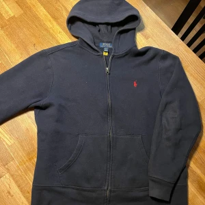 Marinblå hoodie från Ralph Lauren - Säljer en snygg och bekväm marinblå hoodie från Ralph Lauren. Den har en dragkedja framtill och en klassisk röd logga på bröstet. Perfekt för både höst och vinter, och den är i bra skick. Passar till alla tillfällen!