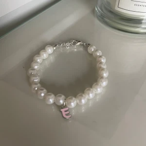 Pärlarmband med bokstavsberlock - Säljer ett superfint armband med vita pärlor och en söt rosa bokstavsberlock 'E'. Perfekt för att ge en personlig touch till din outfit. Armbandet har en enkel och elegant design som passar både till vardags och fest. 