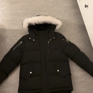 Svart dunjacka från Moose Knuckles - HELT NY! Säljer en snygg och varm svart dunjacka från Moose Knuckles. Jackan har en fluffig vit pälsdetalj runt huvan och är perfekt för kalla vinterdagar. Den har långa ärmar och praktiska fickor med dragkedja och knappar. 