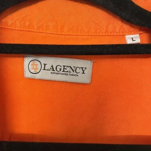 Orange kortärmad skjorta från Lagency - Säljer en snygg orange kortärmad skjorta från Lagency i storlek L. Skjortan är tillverkad i 100% Tencel vilket gör den superbekväm och perfekt för sommardagar. Den har knappar hela vägen ner och en klassisk krage. Perfekt för både vardag och fest! 🍊