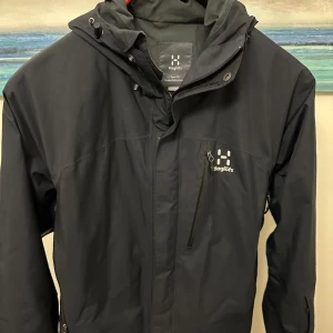 Svart vindjacka från Haglöfs - Säljer en svart regnjacka från Haglöfs i storlek M. Jackan har en praktisk dragkedja och en justerbar huva. Perfekt för blåsiga dagar och utomhusaktiviteter. Den är i bra skick!