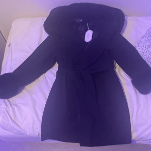 Svart kappa med pälsdetaljer - Säljer en elegant svart kappa med fluffig päls runt huvan och ärmsluten. Den har ett bälte i midjan för en snygg passform. Perfekt för kyliga höst- och vinterdagar när du vill hålla dig varm och stilren.