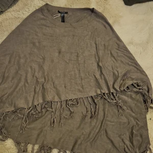 Beige/brun poncho från Comma - Säljer en superfin beige/ brunponcho från Comma. Den är i ett mjukt material och har fransar längs nederkanten som ger en bohemisk touch. Perfekt för höst och vår när du vill ha något lätt men ändå stiligt. Passar bra över en långärmad tröja eller blus.