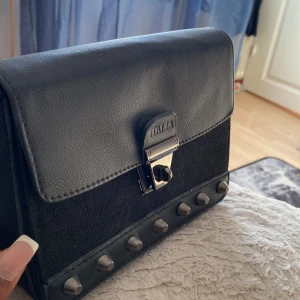 Snygg svart handväska  - Det här är en skitsnygg handväska med fräcka detaljer. I väskan finns även en liten ficka med dragkedja, där man kan lägga in kort osv. Det finns även ett snöre som man kan sätta fast på väskan och ha det runt axeln. Har knappt använt den!