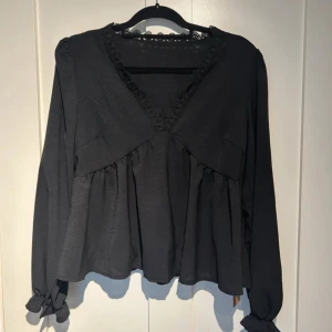 Svart blus med spetsdetaljer - Säljer en elegant svart blus med spetsdetaljer vid halsringningen. Blusen har långa ärmar med knytband vid ärmsluten och en v-ringad design. Perfekt för höst och vinter, både till vardags och festliga tillfällen.