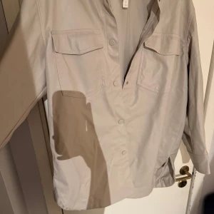 Beige läderjacka från H&M - Snygg beige jacka från H&M med knappar framtill och två bröstfickor. Perfekt för en stilren look. Skjortan har långa ärmar och en klassisk krage. Endast använd 2-3 gånger.