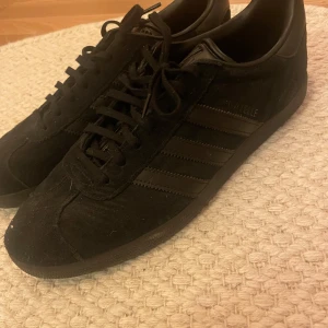 Svarta Adidas Gazelle sneakers - Snygga svarta Adidas Gazelle sneakers i mocka med klassiska tre ränder på sidan. Skorna har snörning och en stilren design som passar perfekt till vardags.