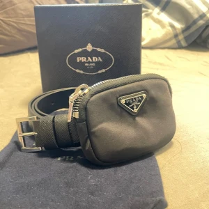 Svart bälte med väska från Prada - Snyggt svart bälte från Prada med en liten matchande väska. Bältet har en elegant silverfärgad spänne med Prada-logotypen. Perfekt för att bära småsaker med stil. Size 90