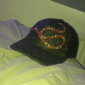 Svart keps med ormmönster - Snygg svart keps från Gucci med ett ormmönster i rött och vitt. Kepsen har ett justerbart spänne baktill och är tillverkad i ett slitstarkt material med märkets ikoniska mönster. Box finns ej länge men pgrund av en flytt men kan garantera att den e äkta