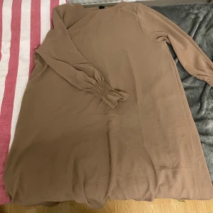 Beige abaya klänning - Säljer en stilren beige abaya med långa ärmar och ribbade detaljer vid ärmsluten. Perfekt för en avslappnad och elegant look.
